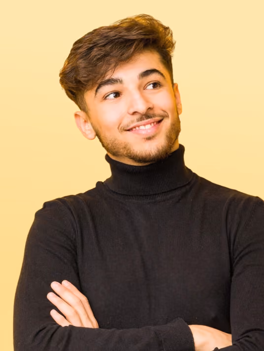 Young man with brown hair and beard wearing a black turtleneck sweater, smiling and looking to the side with arms crossed against a yellow background.