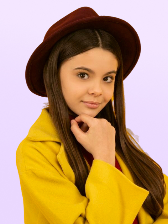 Young woman wearing a brown hat and yellow coat, posing with her hand near her chin against a light purple background.