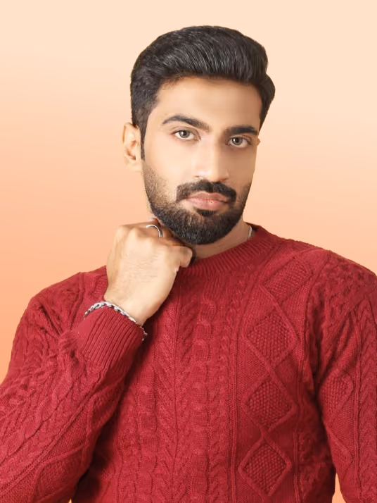 Man with dark hair and beard wearing a red cable-knit sweater, posing with his fist near his chin against a peach-colored background.