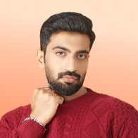 Man with dark hair and beard wearing a red sweater, resting his chin on his hand against a peach background.