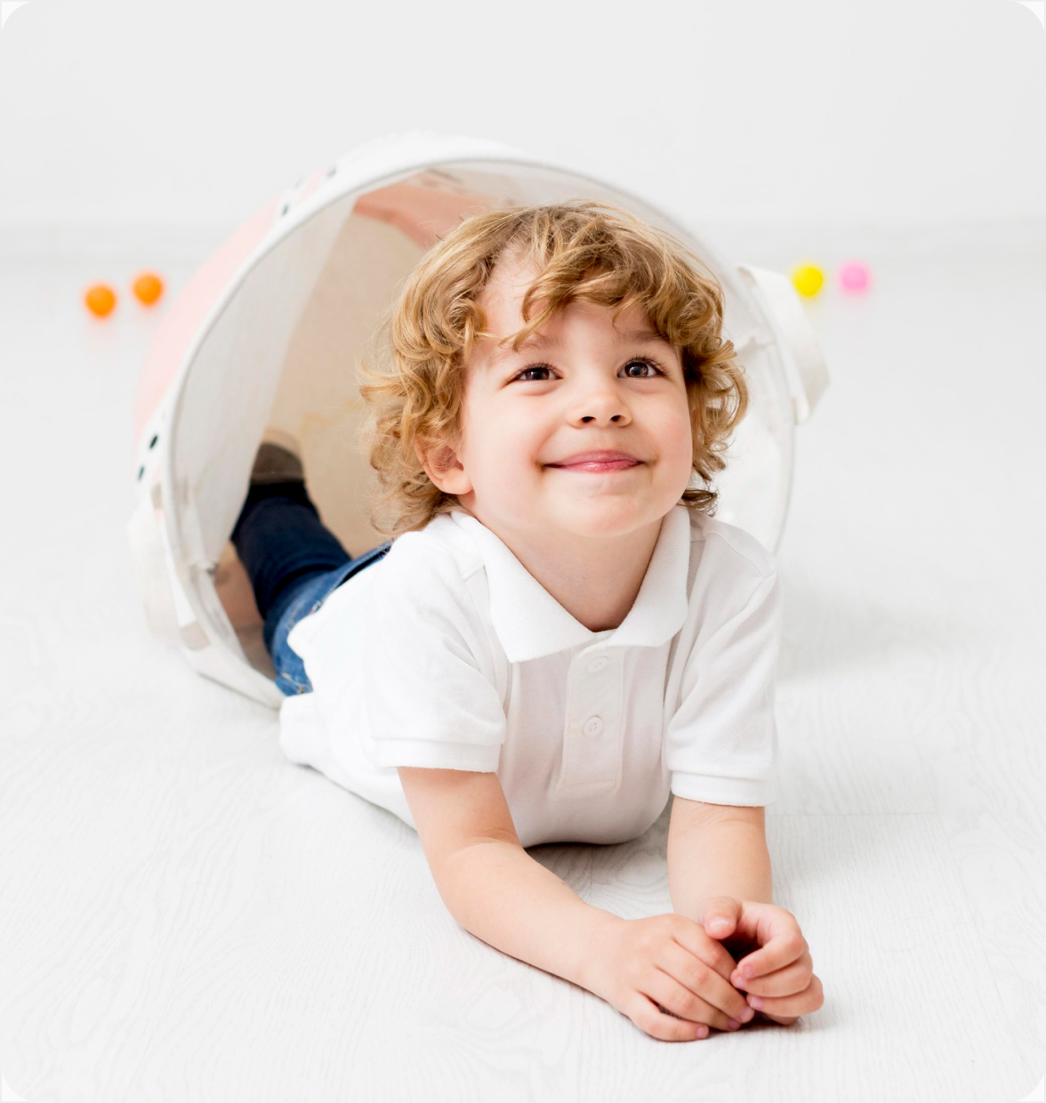 Smiling young boy with curly hair lying on floor inside a large toy bucket.