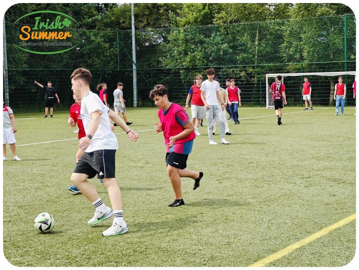 Jugendliche spielen Fußball auf einem grünen Sportplatz mit Tor im Hintergrund.