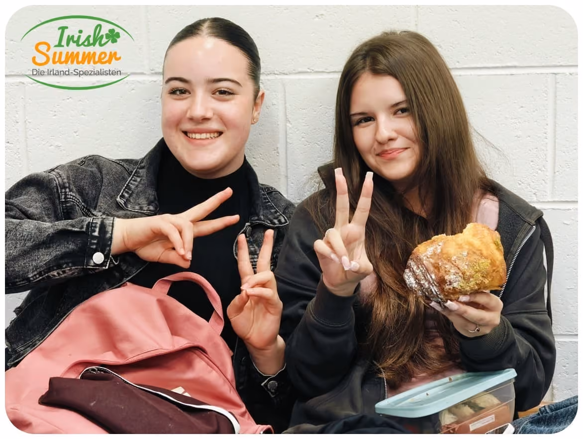 Zwei junge Frauen sitzen vor einer weißen Wand, lächeln und zeigen mit den Fingern Frieden, eine hält ein Gebäckstück.