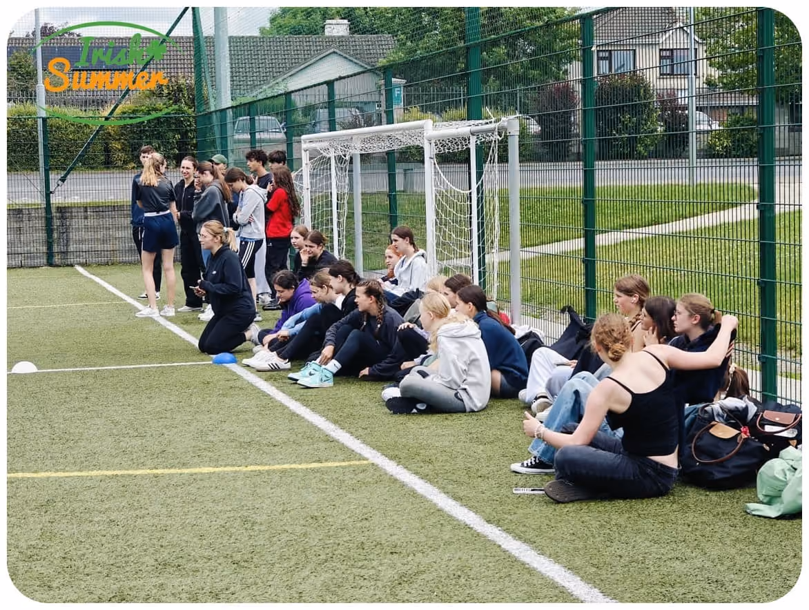 Gruppe Jugendlicher sitzt und steht auf einem Sportplatz neben einem Fußballtor, umgeben von Zaun und Häusern.