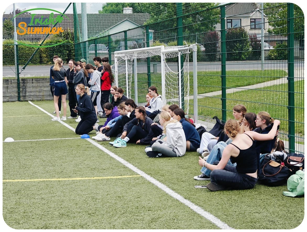 Gruppe Jugendlicher sitzt und steht auf einem Sportplatz neben einem Fußballtor, umgeben von Zaun und Häusern.