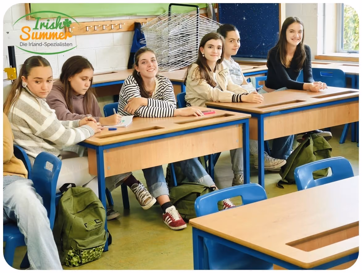 Sechs Schülerinnen sitzen an Holztischen mit blauen Stühlen in einem hellen Klassenzimmer.