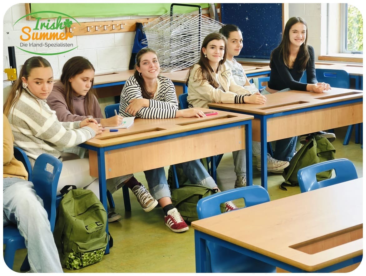 Sechs Schülerinnen sitzen an Holztischen mit blauen Stühlen in einem hellen Klassenzimmer.
