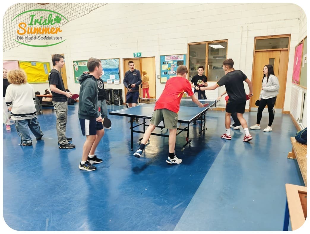 Jugendliche spielen Tischtennis in einer Turnhalle mit blauem Boden und beigen Wänden.
