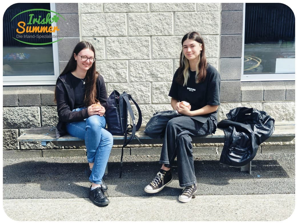 Zwei junge Frauen sitzen auf einer Holzbank vor einer Steinfassade, beide halten ein Brötchen in den Händen, eine hat eine schwarze Jacke neben sich.