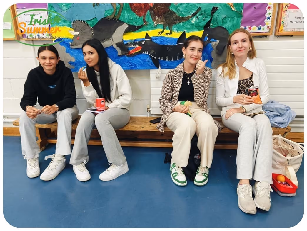 Vier junge Frauen sitzen auf einer Holzbühne vor einer Wand mit einer bunten Tiermalerei.