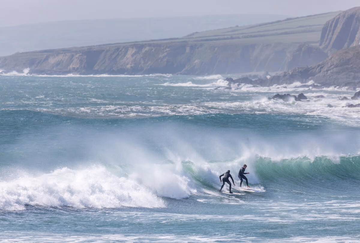 Zwei Surfer in Neoprenanzügen reiten auf einer Welle am Küstenabschnitt mit Klippen im Hintergrund.