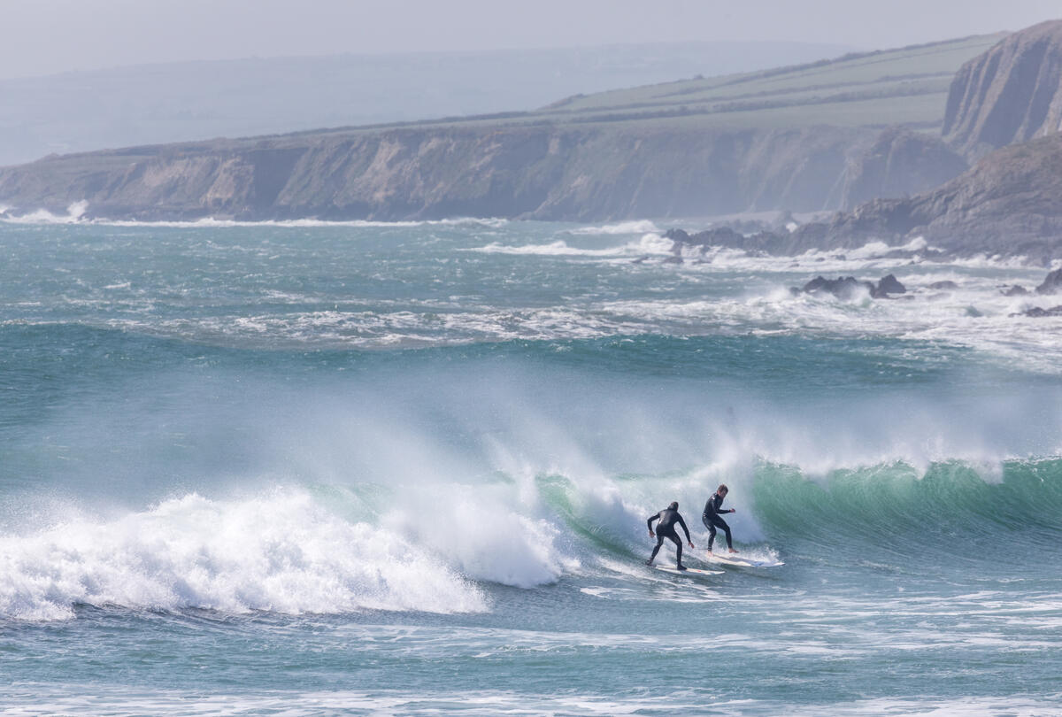 Zwei Surfer in Neoprenanzügen reiten auf einer Welle am Küstenabschnitt mit Klippen im Hintergrund.