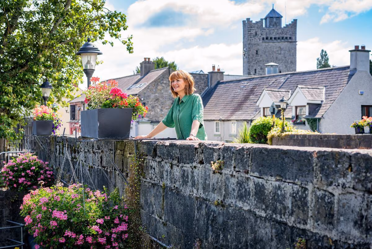 Frau in grünem Hemd lehnt lächelnd an einer Steinmauer mit Blumen in Töpfen, im Hintergrund Steinhaus und Turm bei sonnigem Himmel.