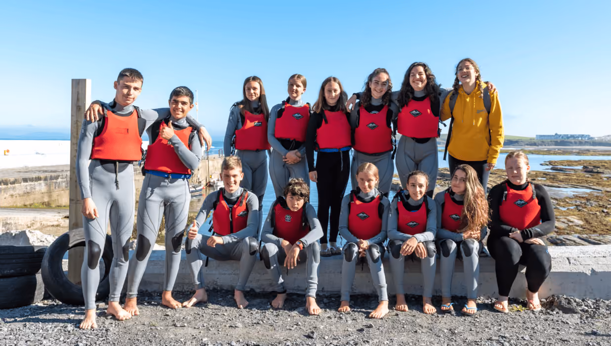 Gruppenfoto von Jugendlichen in Neoprenanzügen mit roten Westen am Meeresufer unter klarem blauem Himmel.