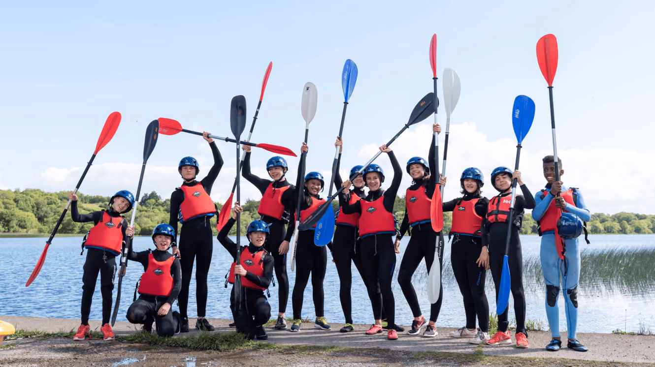 Gruppe von Jugendlichen in roten Schwimmwesten und Helmen hält bunte Kajakpaddel am Seeufer hoch.