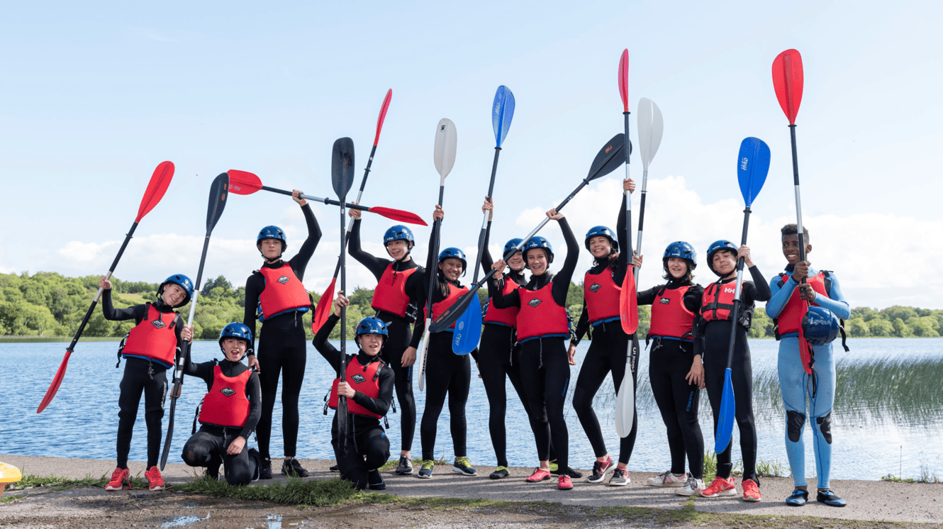 Gruppe von Jugendlichen in roten Schwimmwesten und Helmen hält bunte Kajakpaddel am Seeufer hoch.