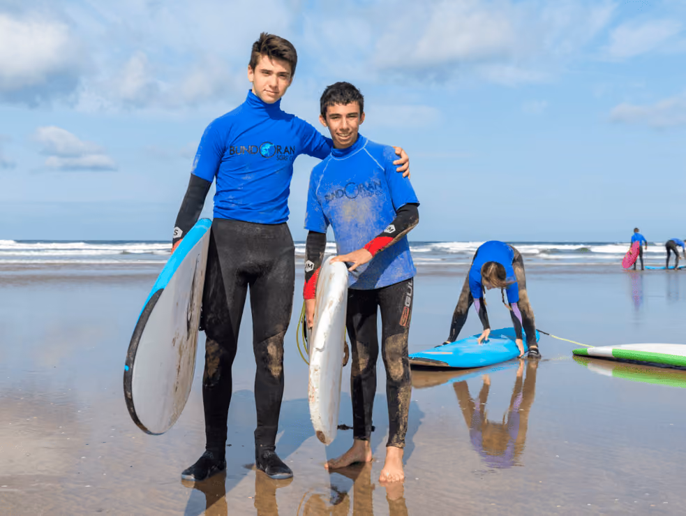 Zwei Jugendliche in blauen Surfanzügen stehen am Strand und halten Surfbretter, während andere im Hintergrund Surfbretter vorbereiten.