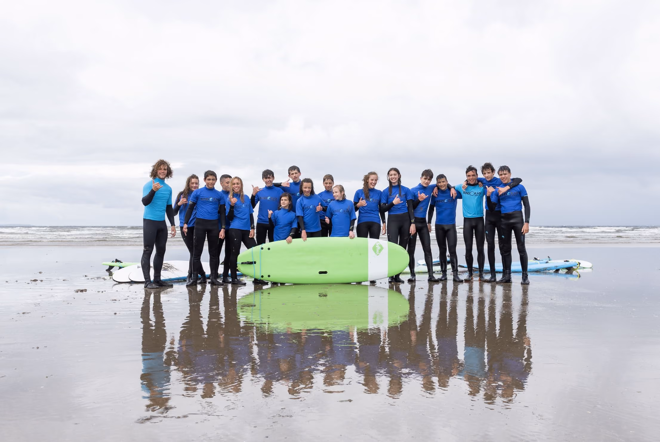 Gruppe von Jugendlichen in blauen Neoprenanzügen posiert mit Surfbrettern am Strand unter bewölktem Himmel.