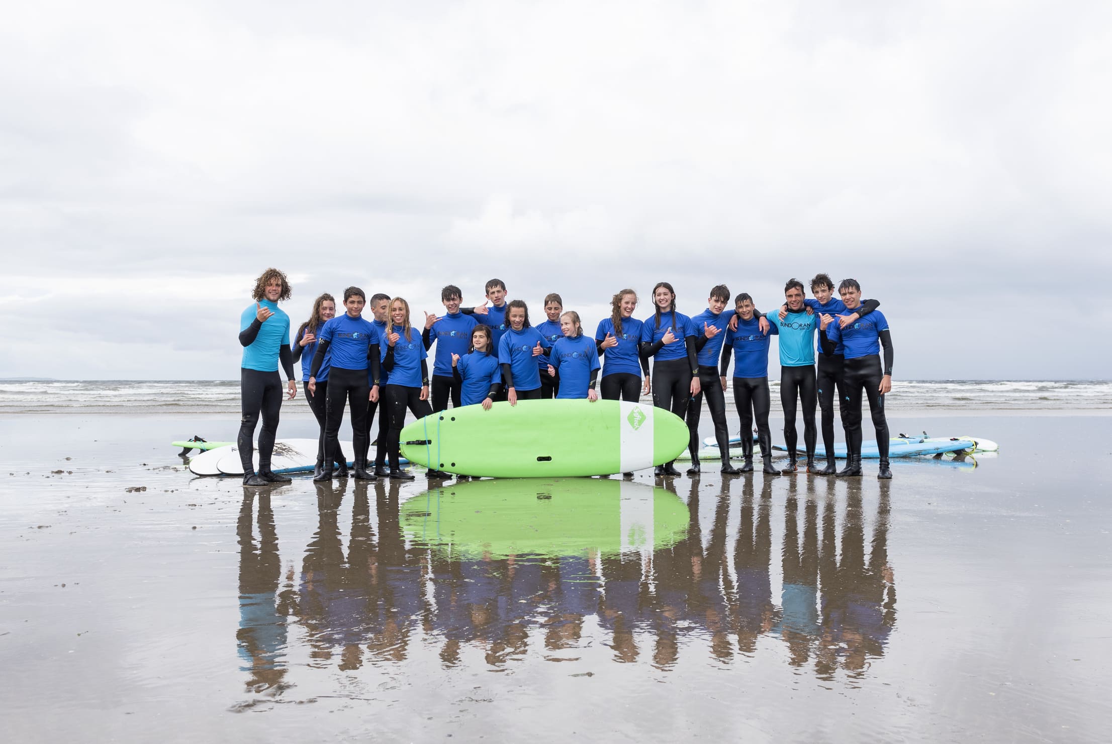 Gruppe von Jugendlichen in blauen Neoprenanzügen posiert mit Surfbrettern am Strand unter bewölktem Himmel.