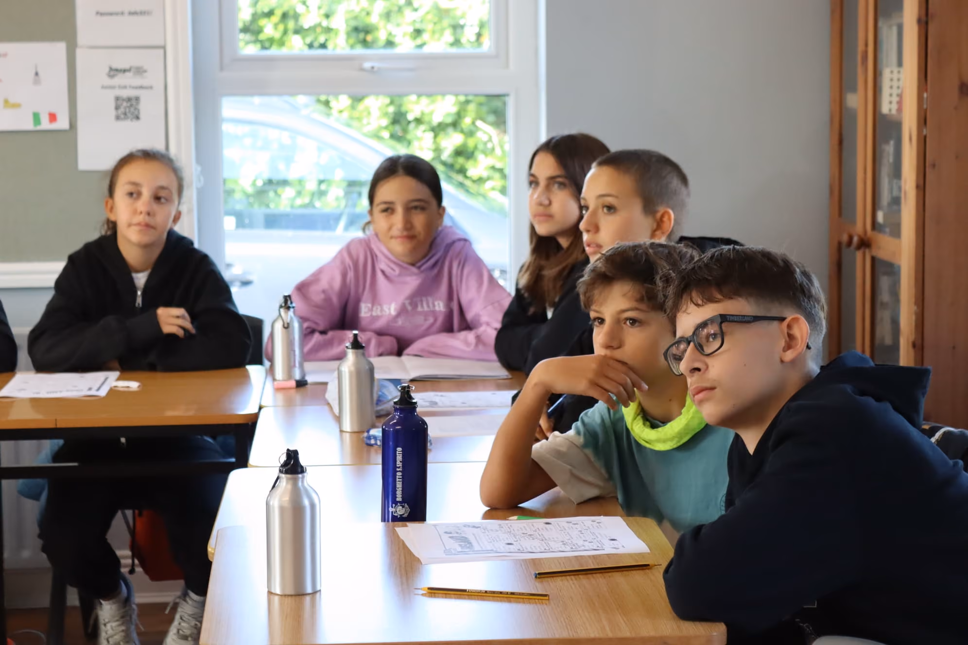 Sechs Jugendliche sitzen an Schultischen in einem Klassenzimmer und hören aufmerksam zu.