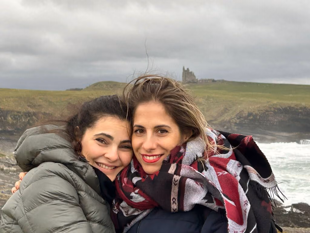 Zwei Frauen in Winterbekleidung umarmen sich lächelnd vor einer Küstenlandschaft mit grünem Gras und einer Burgruine im Hintergrund unter bewölktem Himmel.