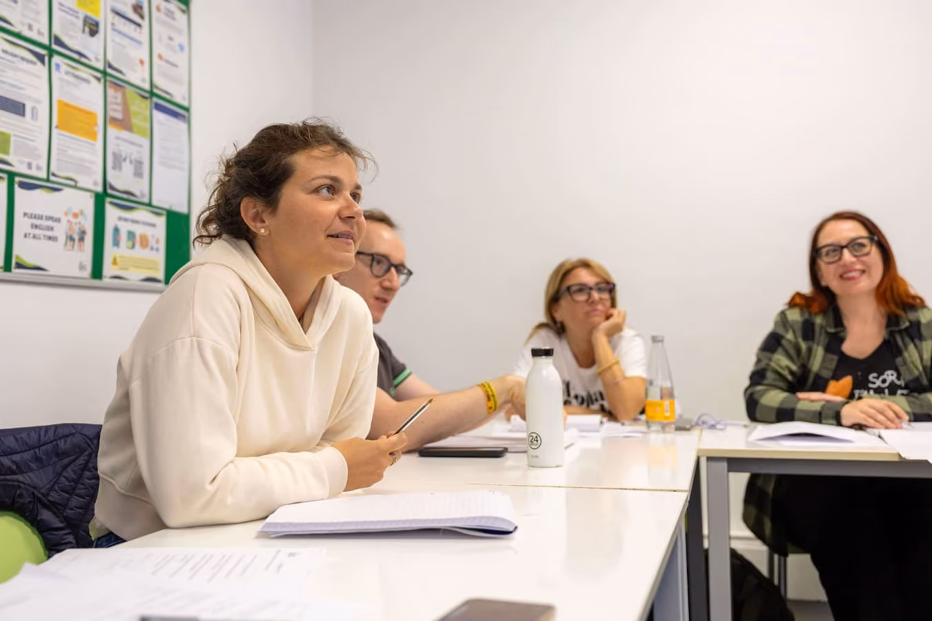 Vier Erwachsene sitzen in einem Klassenzimmer an weißen Tischen und hören aufmerksam zu.