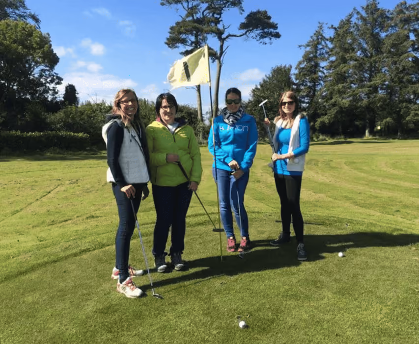 Vier Frauen stehen auf einem grünen Golfplatz neben Fahne und Golfbällen, halten Golfschläger bei sonnigem Himmel.