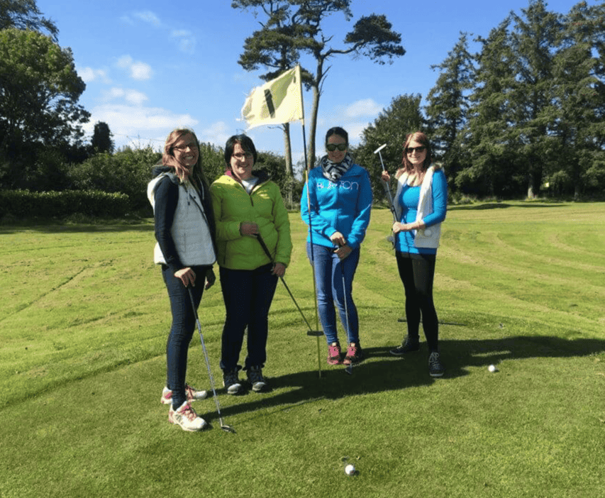 Vier Frauen stehen auf einem grünen Golfplatz neben Fahne und Golfbällen, halten Golfschläger bei sonnigem Himmel.