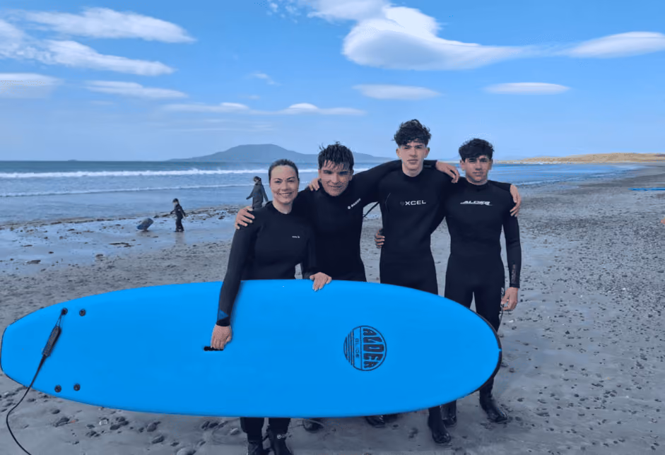 Vier Personen in Neoprenanzügen stehen am Strand, halten ein blaues Surfbrett, mit Meer und Hügel im Hintergrund.
