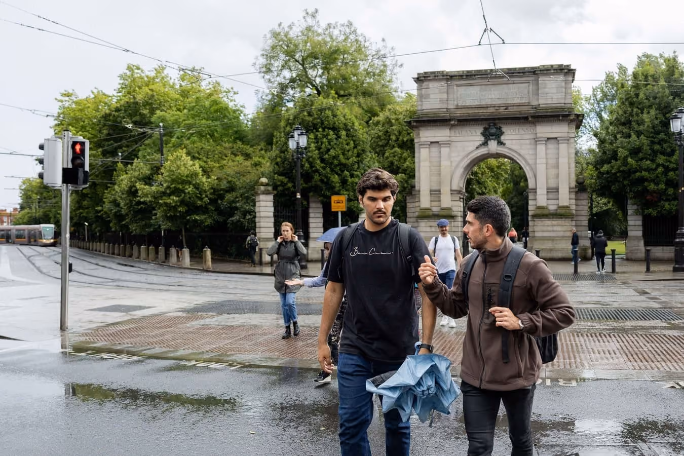 Zwei junge Männer mit Rucksäcken und zusammengefaltetem Regenschirm in der Hand gehen auf einer nassen Straße vor einem steinernen Torbogen, im Hintergrund Bäume und weitere Fußgänger.