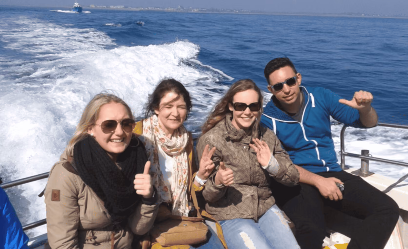 Vier Personen mit Sonnenbrillen sitzen lächelnd auf einem Boot, das über das blaue Meer fährt.