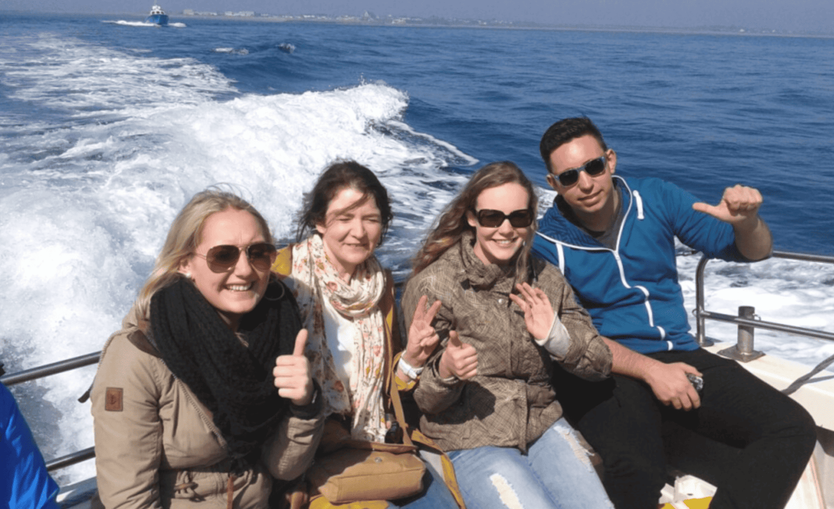 Vier Personen mit Sonnenbrillen sitzen lächelnd auf einem Boot, das über das blaue Meer fährt.