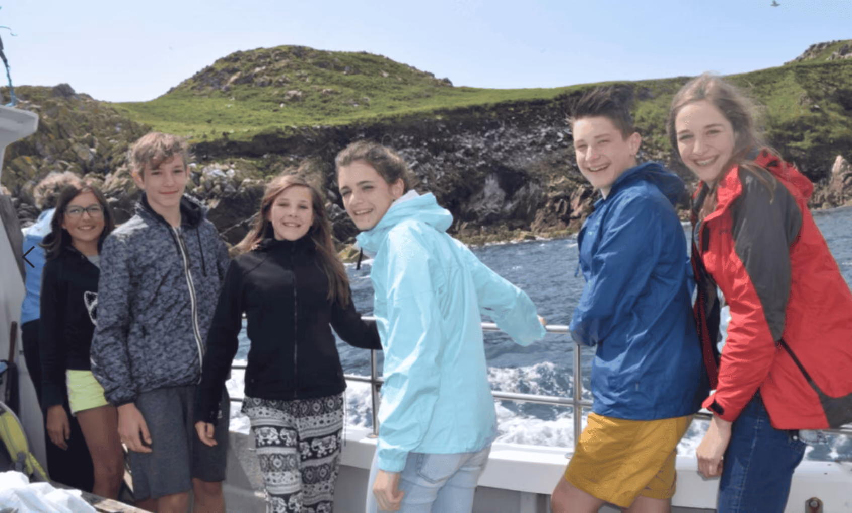 Sechs Jugendliche in leichten Jacken posieren lächelnd auf einem Boot vor grünen, felsigen Hügeln und Meer.