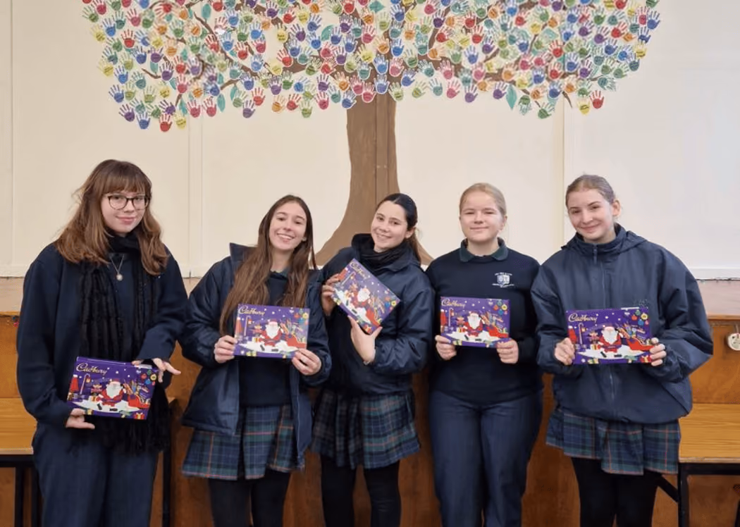 Fünf Mädchen in Schuluniformen stehen vor einer Wand mit einem Baum aus bunten Handabdrücken und halten Cadbury-Pralinenboxen.