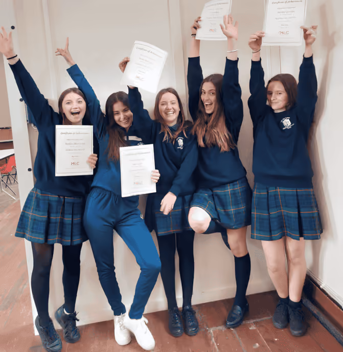 Fünf glückliche Schülerinnen in Uniformen halten ihre Zertifikate hoch und jubeln in einem Klassenzimmer.