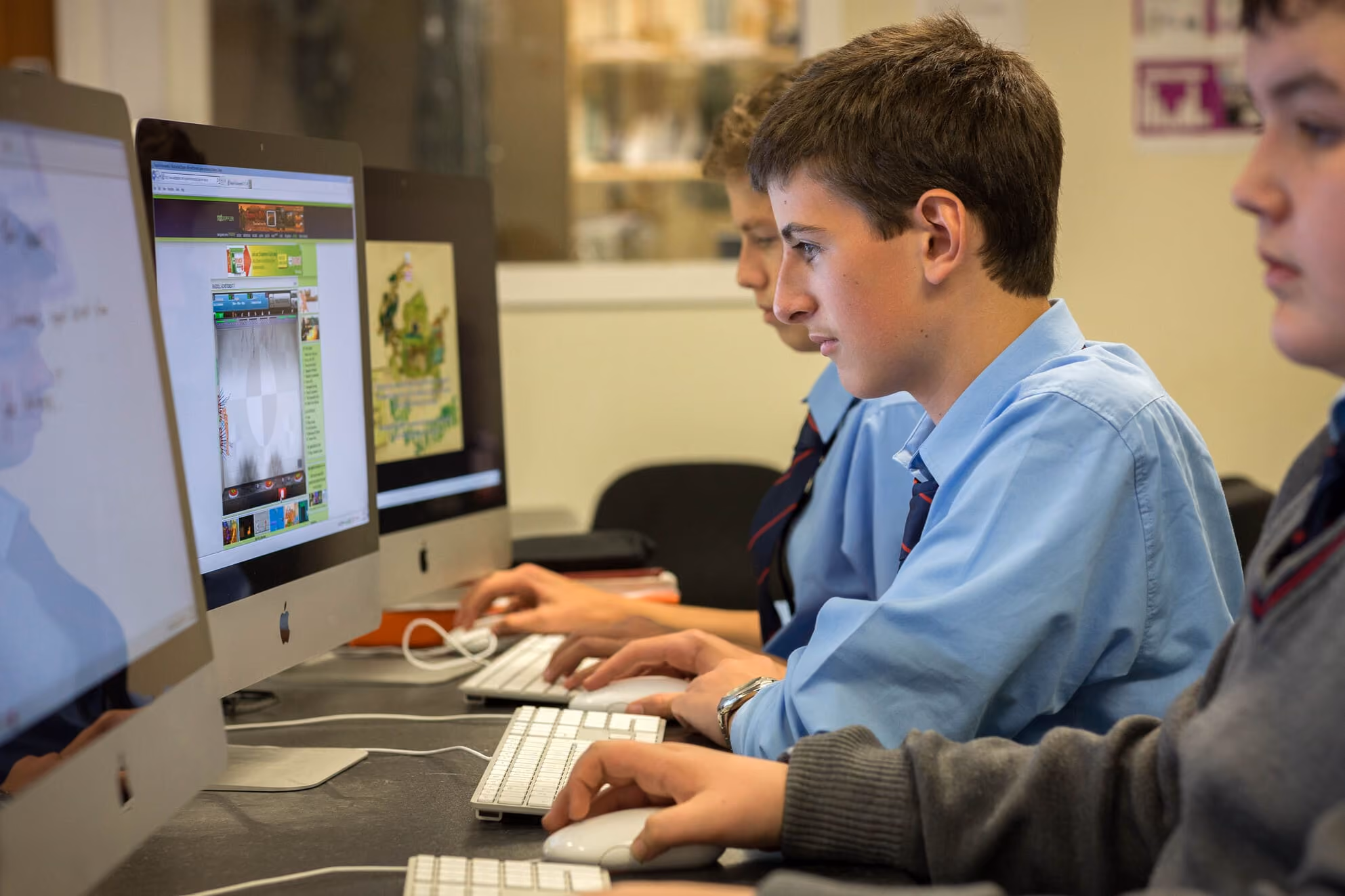 Drei Schüler in Schuluniform sitzen nebeneinander an einem Tisch und arbeiten konzentriert an iMac-Computern.