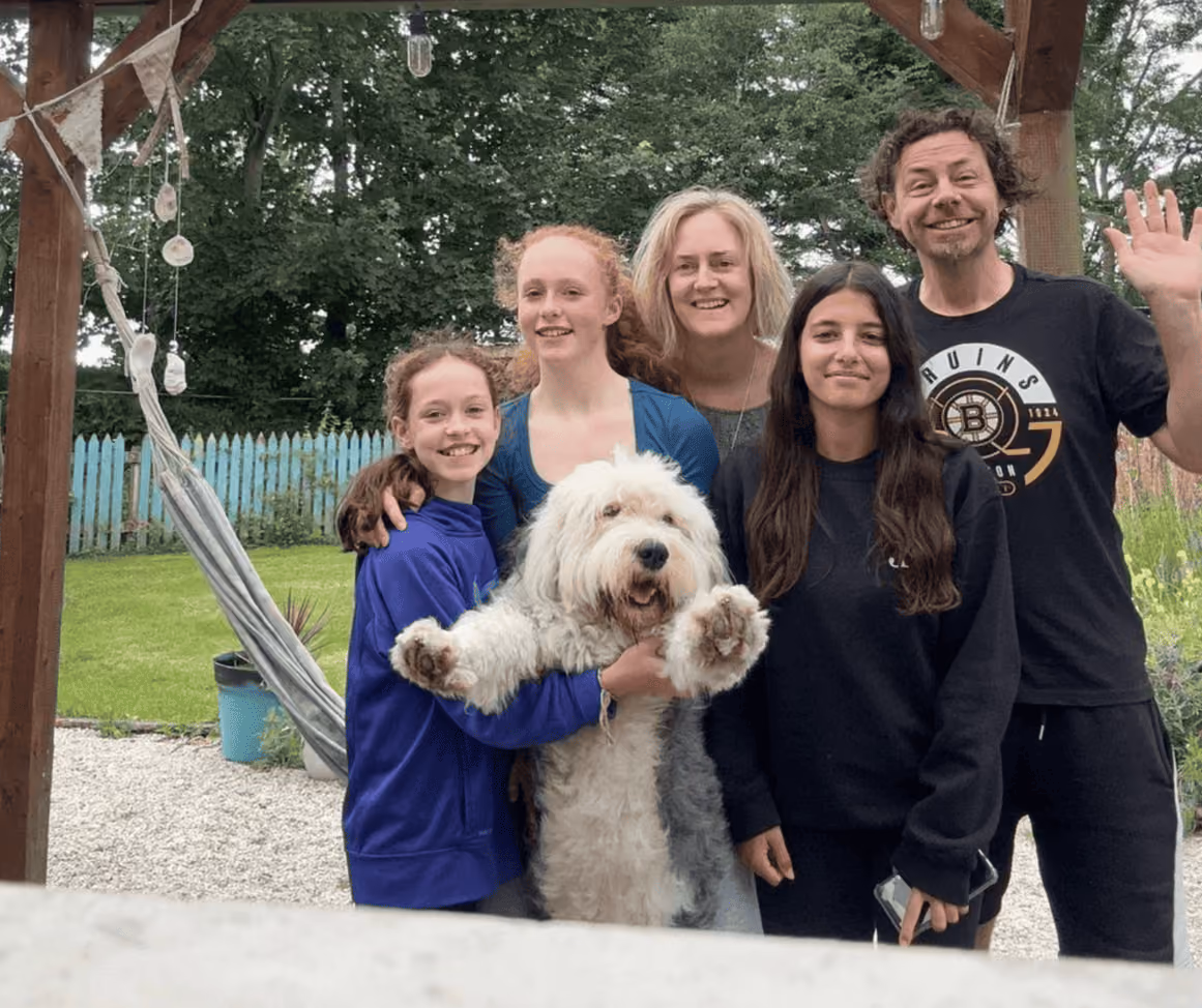 Fünf lächelnde Menschen, darunter zwei Mädchen, eine junge Frau, ein Mann, und ein großer weißer Hund, der mit ausgestreckten Pfoten gehalten wird, vor einem Gartenhäuschen mit grünem Hintergrund.