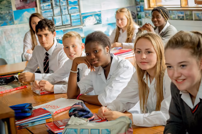 Schüler verschiedener Herkunft sitzen in Schuluniformen in einem Klassenzimmer an einem Holztisch mit Schulbüchern und Schulmaterialien.