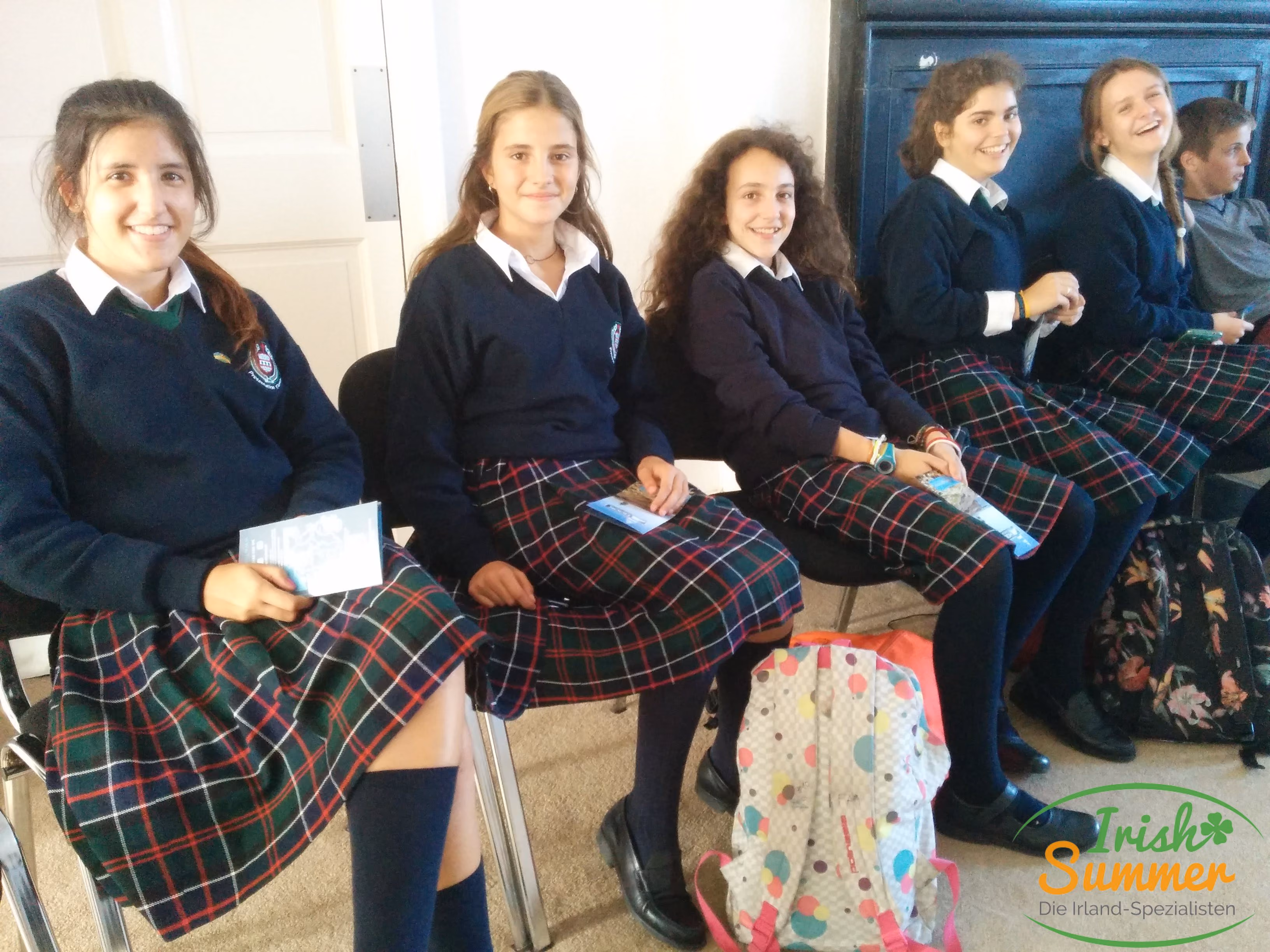 Fünf Mädchen in Schuluniform mit karierten Röcken sitzen auf Stühlen und lächeln, mit zwei Rucksäcken auf dem Boden neben ihnen.