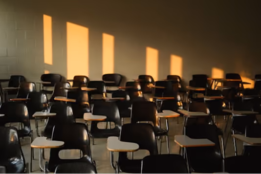 An empty classroom with rows of dark student desk-chairs arranged on a tiled floor. Warm golden light streams through unseen windows on the left wall, casting rectangular light and shadow patterns across the cinder block wall. The mood is quiet and atmospheric.