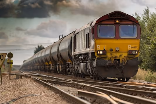 A freight train with a red and yellow locomotive pulls a long line of cylindrical tanker cars along railway tracks in a rural area under a cloudy sky.