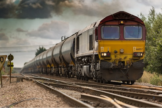 A freight train with a red and yellow locomotive pulls a long line of cylindrical tanker cars along railway tracks in a rural area under a cloudy sky.