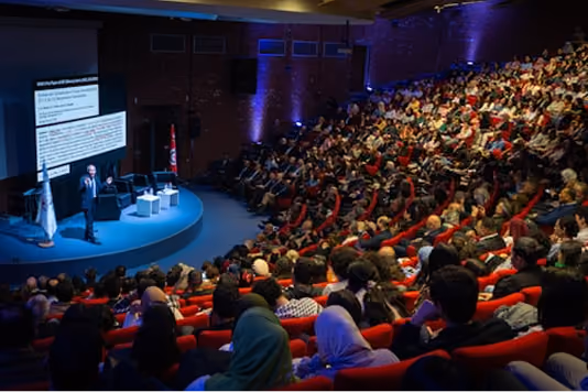 A large lecture hall filled with a diverse audience seated in red chairs, listening to a speaker presenting on stage next to a projected slide. The room is dimly lit with blue accent lighting, and the speaker stands at a podium addressing the crowd.