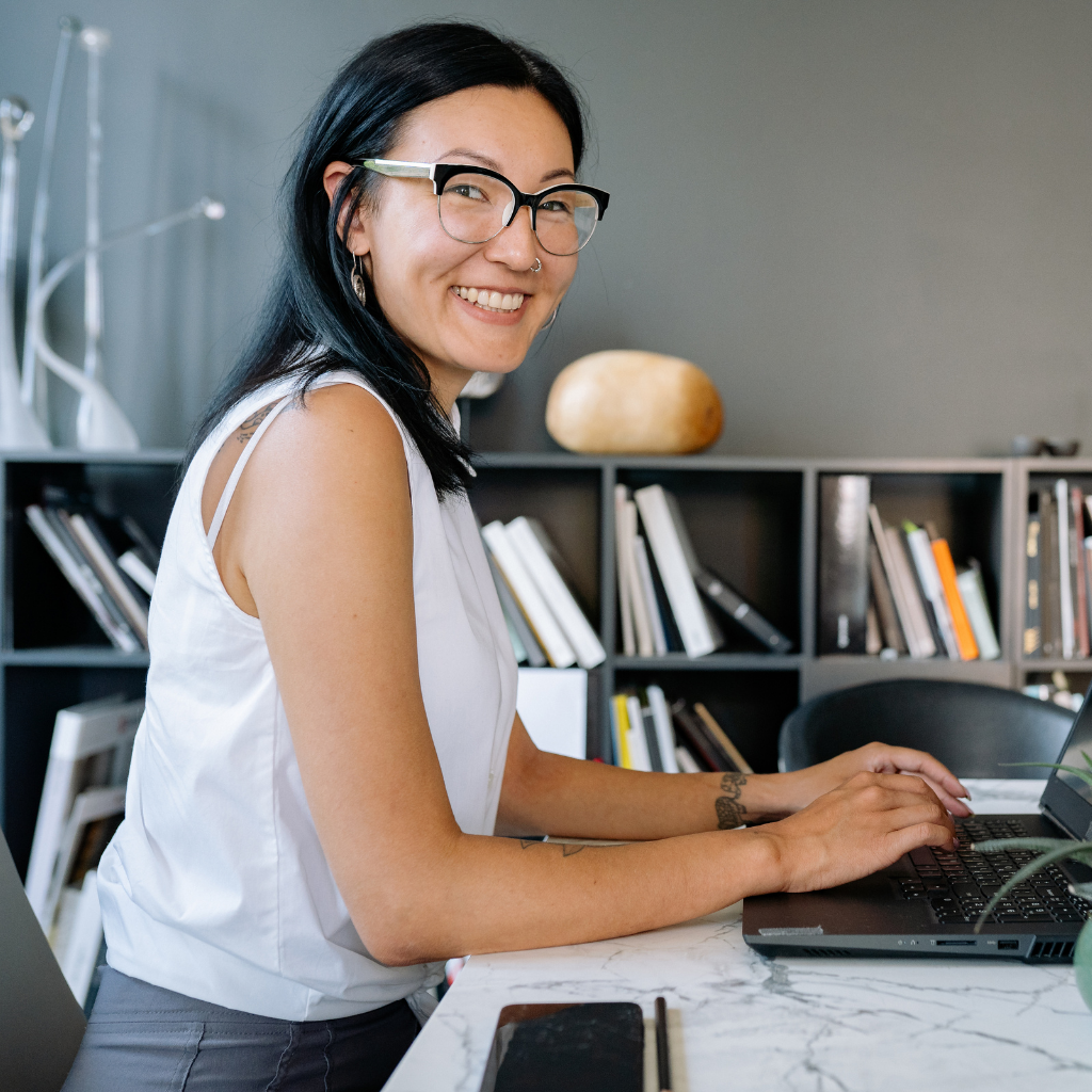 Smiling woman with glasses typing on a laptop at a marble table in a room with a bookshelf background.