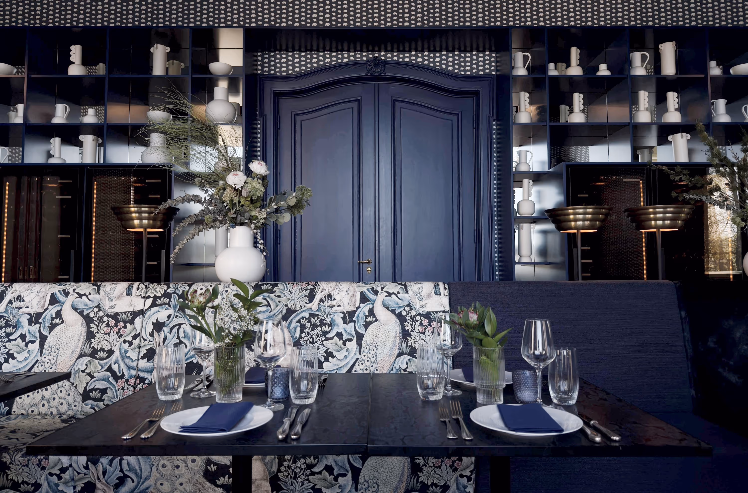Elegantes Restaurant-Interieur mit blauer Doppeltür, botanischem Muster auf der Sitzbank, Tischgedeck mit Gläsern und Vasen mit Blumen.