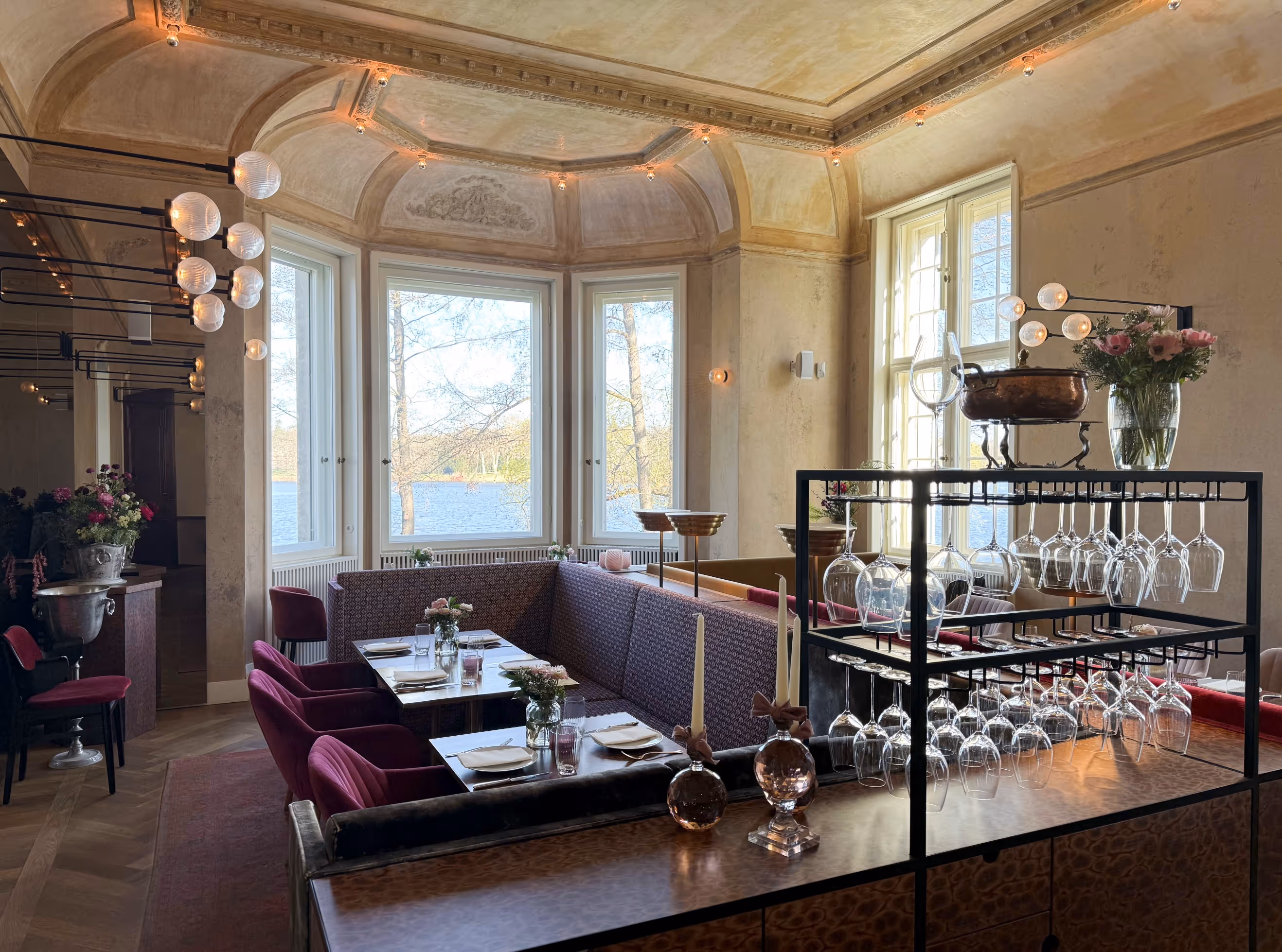Elegantes Restaurantinterieur mit gedeckten Tischen, Samtsesseln, großer Fensterfront mit Seeblick und Hängevorrichtung mit Weingläsern.