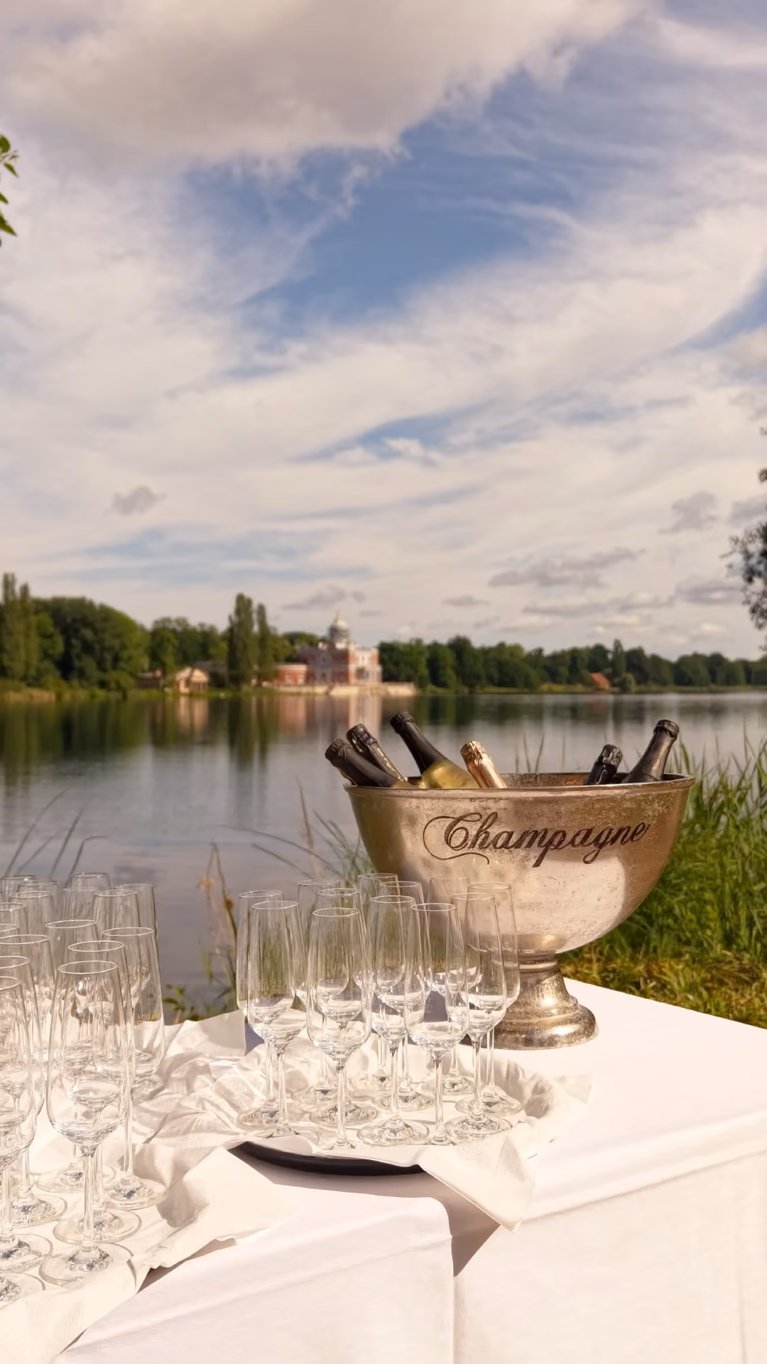 Sektflaschen in einem Eiskübel und leere Gläser auf einem Tisch vor einem See mit Schloss im Hintergrund.