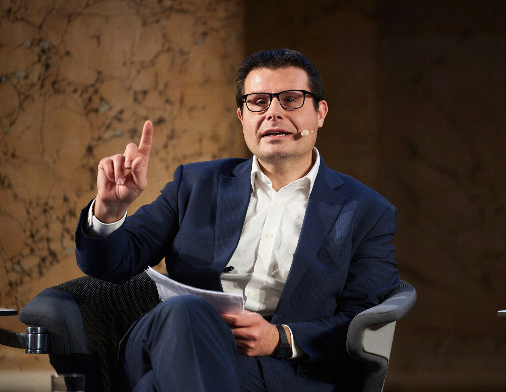 Man in a blue suit and glasses speaking while seated, holding papers and gesturing with one hand raised.