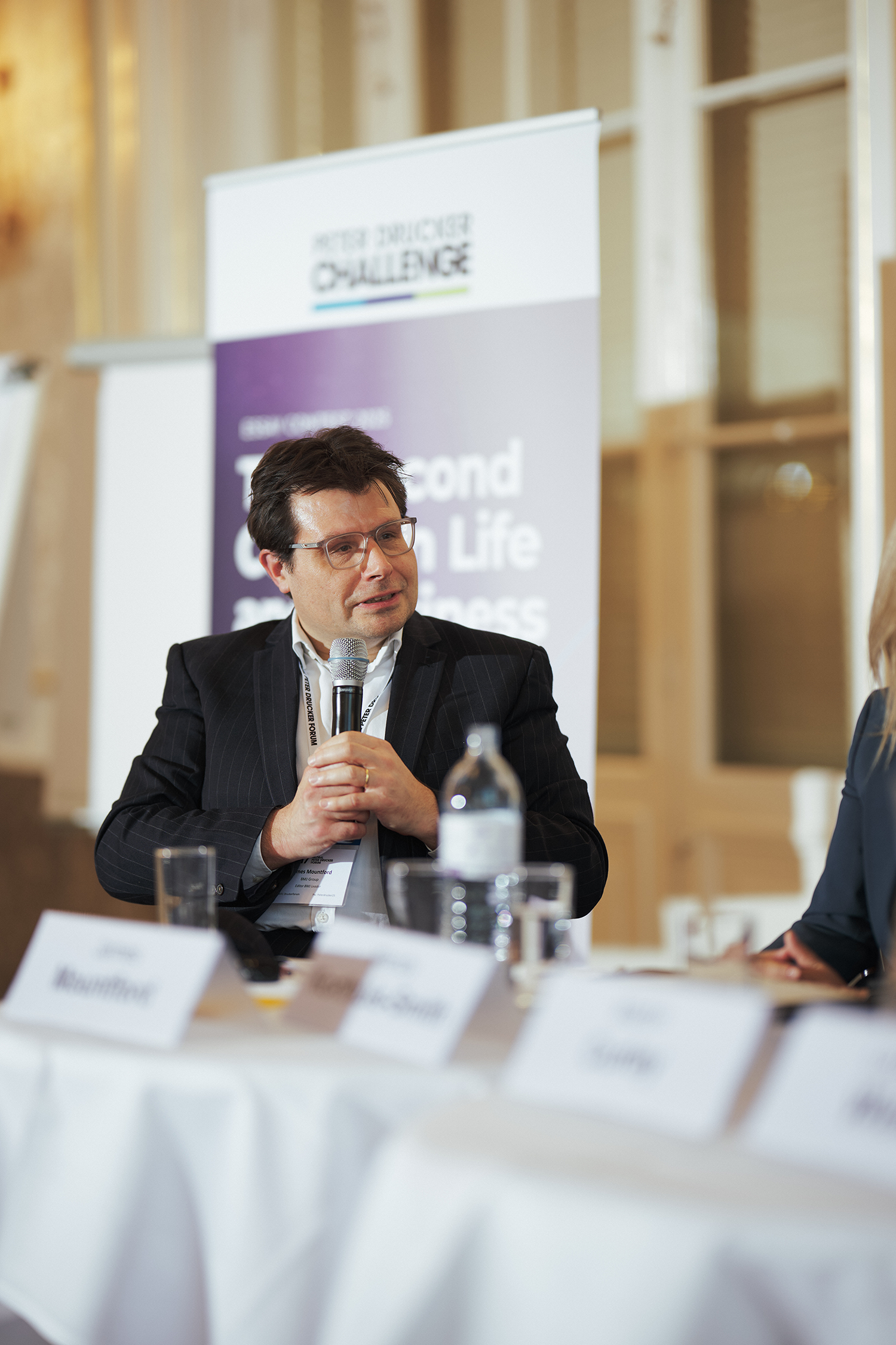 Man in a suit holding a microphone speaking at a panel discussion with a poster in the background reading 'Peter Drucker Challenge'.