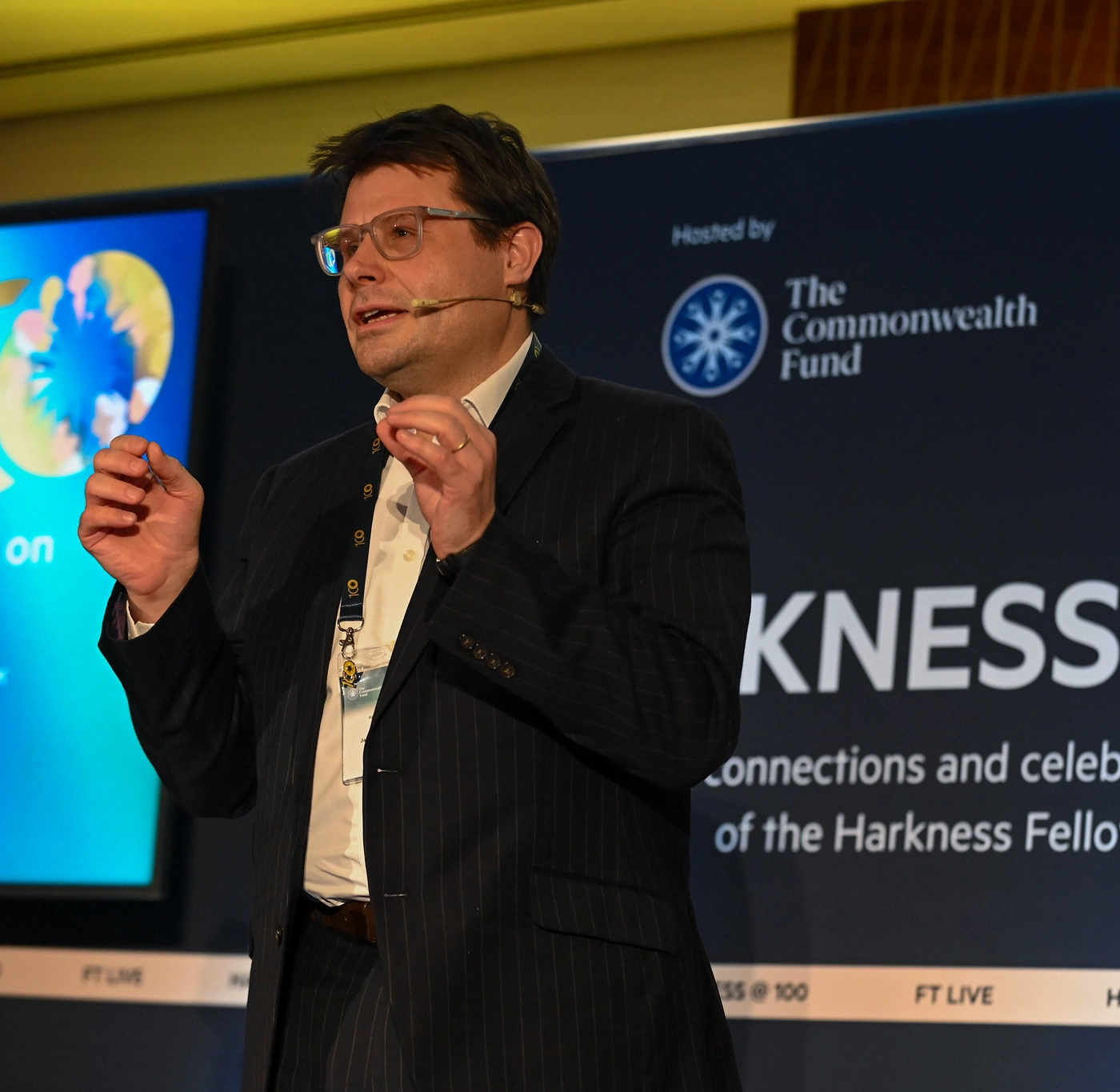 Man in glasses and a dark suit speaking on stage with a microphone headset at a Harkness Fellowship event.